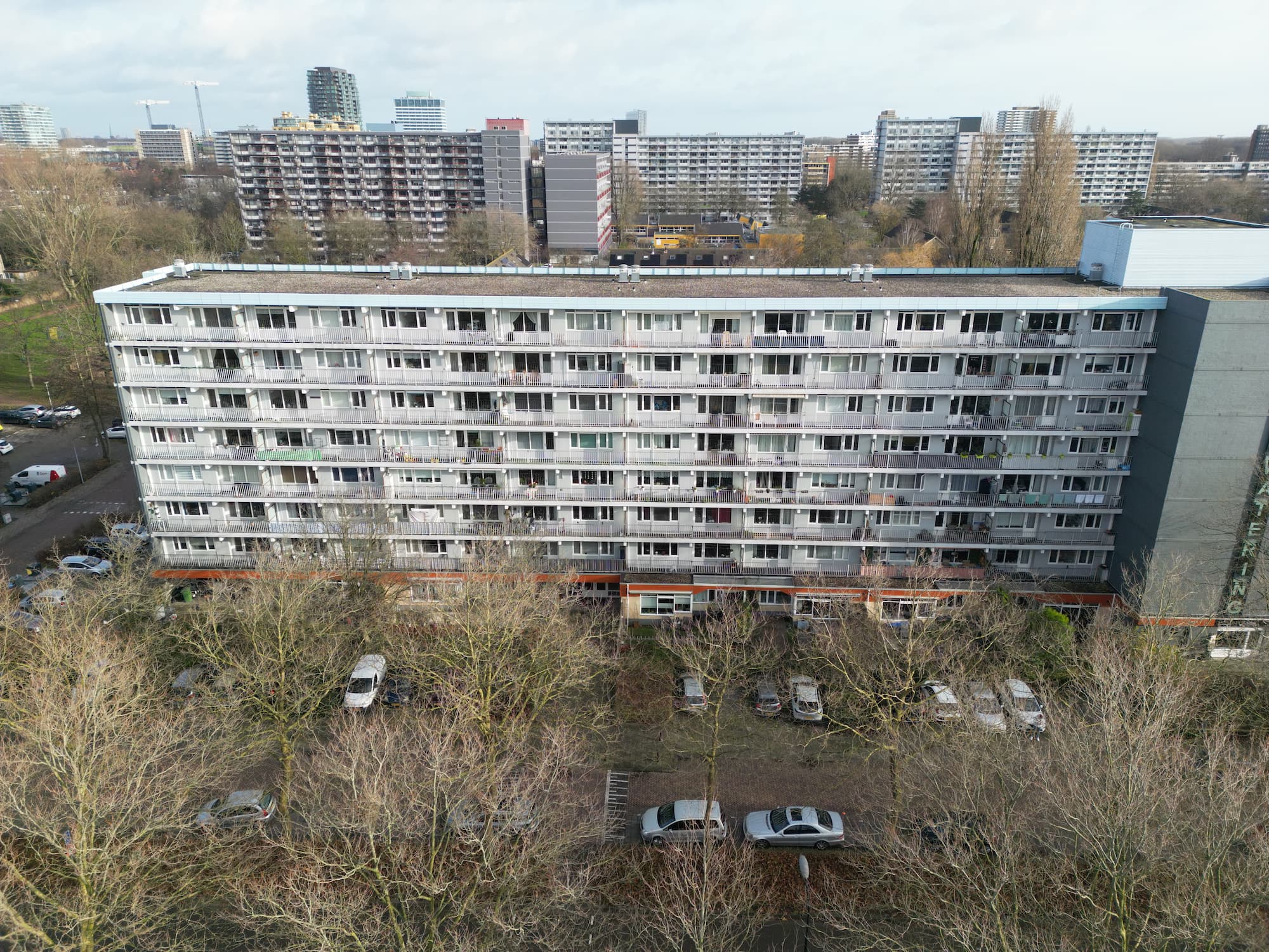 Een vooraanzicht van een hoog appartementsgebouw in een stedelijke omgeving omringd door kale bomen en auto's.