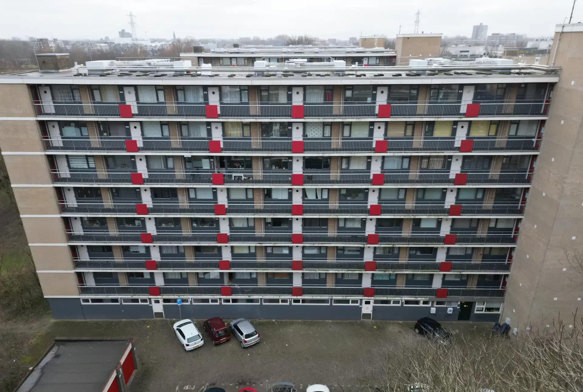 Flatgebouw met rode accenten en balkons, opgenomen van bovenaf op een bewolkte dag.