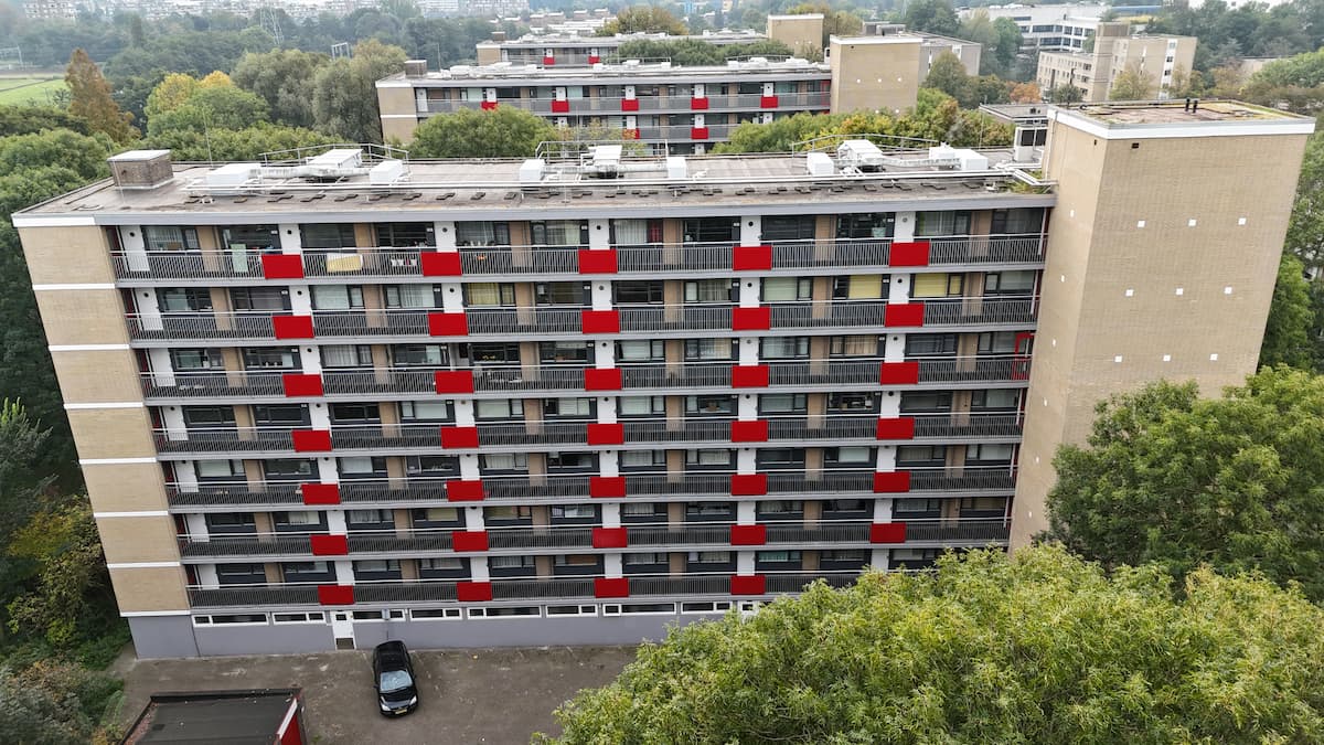 Luchtfoto van een flatgebouw met rode balkonpanelen, omgeven door groen, in een woonwijk.