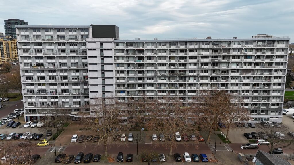 Bovenaanzicht van een groot, grijs wooncomplex met meerdere verdiepingen en een parkeerplaats vol auto's.