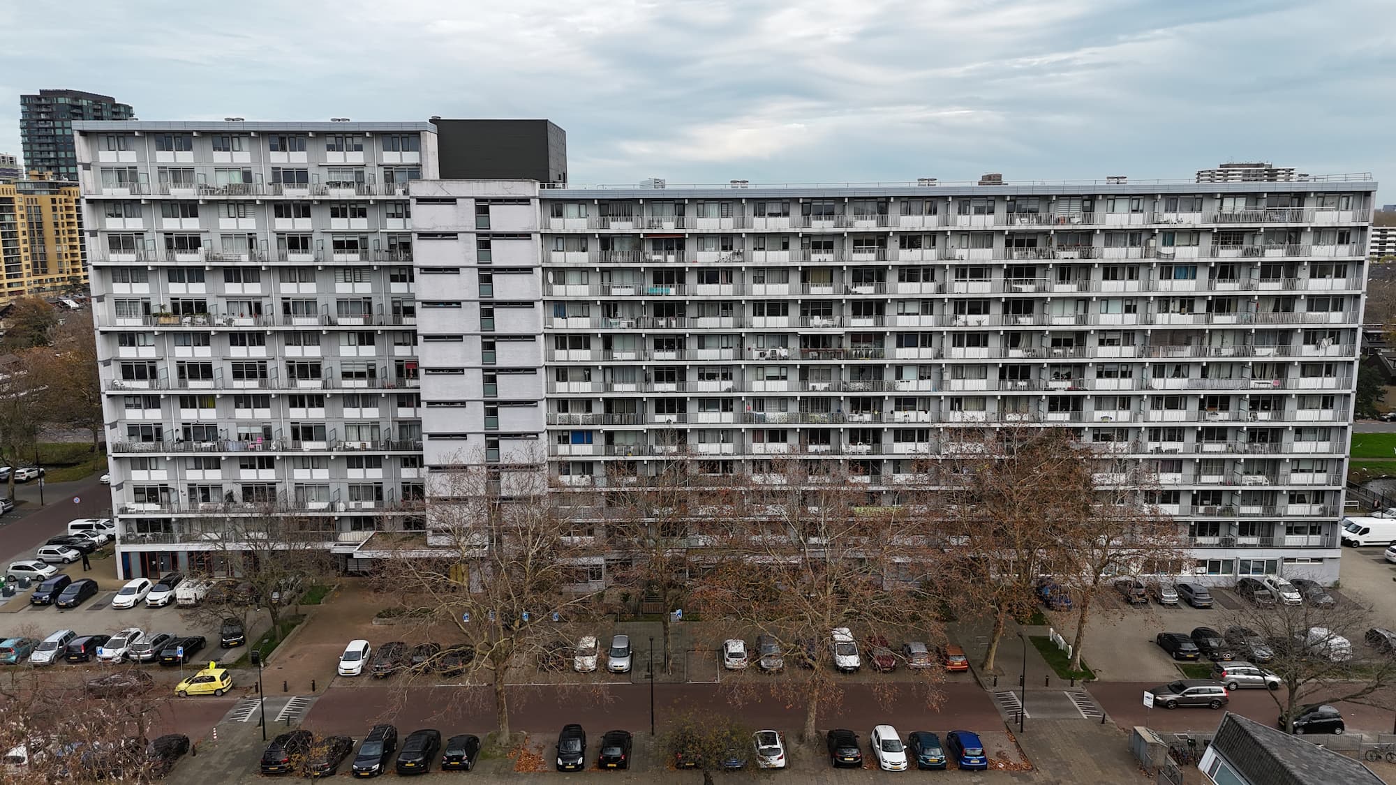 Bovenaanzicht van een groot, grijs wooncomplex met meerdere verdiepingen en een parkeerplaats vol auto's.