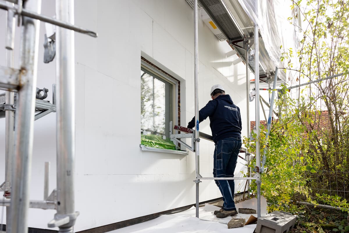 Een bouwvakker is bezig met gevelisolatie naast een raam op een steiger aan de buitenkant van een gebouw.