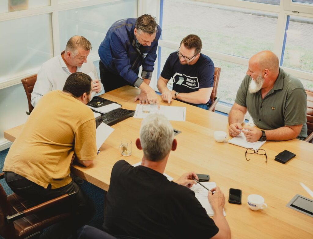 Zes mannen zitten rond een vergadertafel en bespreken documenten tijdens een overleg.