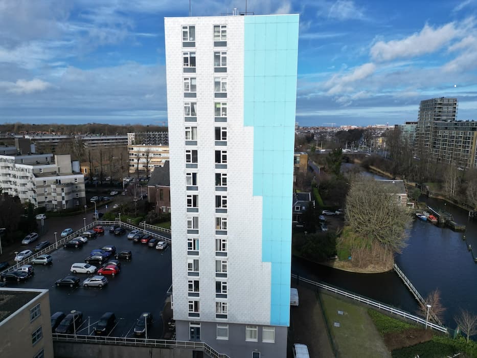 Hoog flatgebouw met blauwe en witte gevel, gelegen naast een parkeerplaats en een kanaal in een stedelijk gebied.