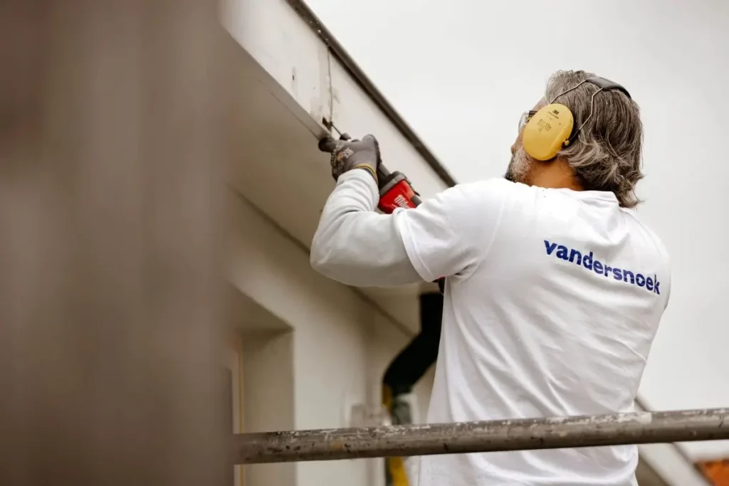 Man in een shirt van Vandersnoek werkt met een elektrisch gereedschap aan het dak van een gebouw.