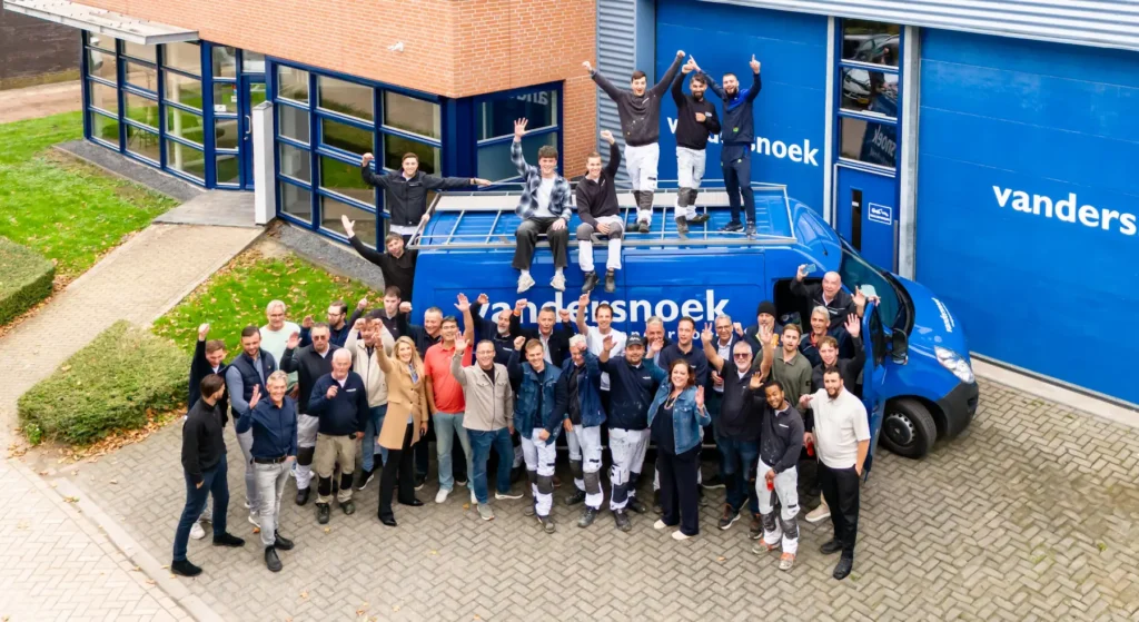 Groepsfoto van het team van Vandersnoek bij hun bedrijfswagen, vrolijk zwaaiend voor hun kantoor.